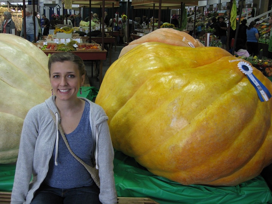 Photo of the author sitting in front of a giant pumpkin that is as big as she is (it has a ribbon it, from a fair).